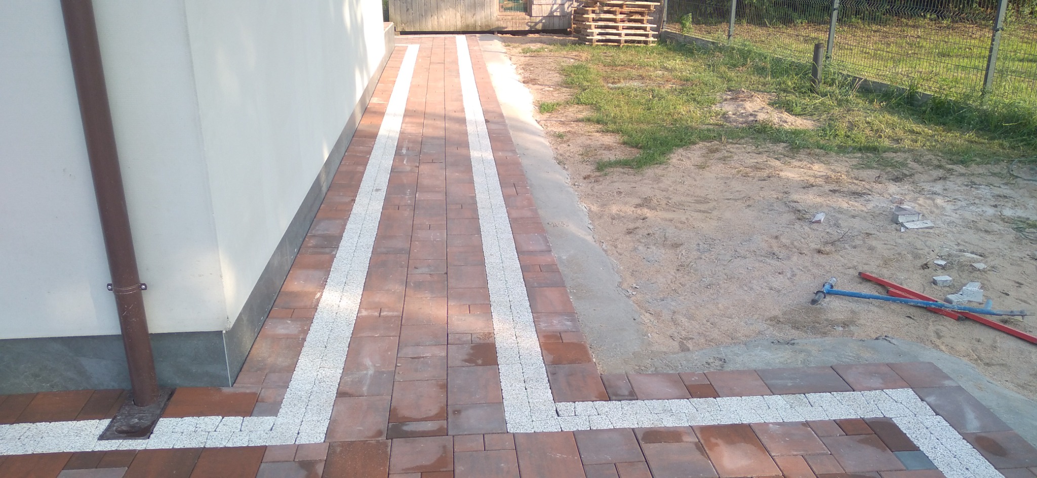 Ułożona kostka brukowa wzdłuż ściany budynku z elementami dekoracyjnymi z jasnego kruszywa, widoczne narzędzia brukarskie i przygotowany teren pod dalsze prace.