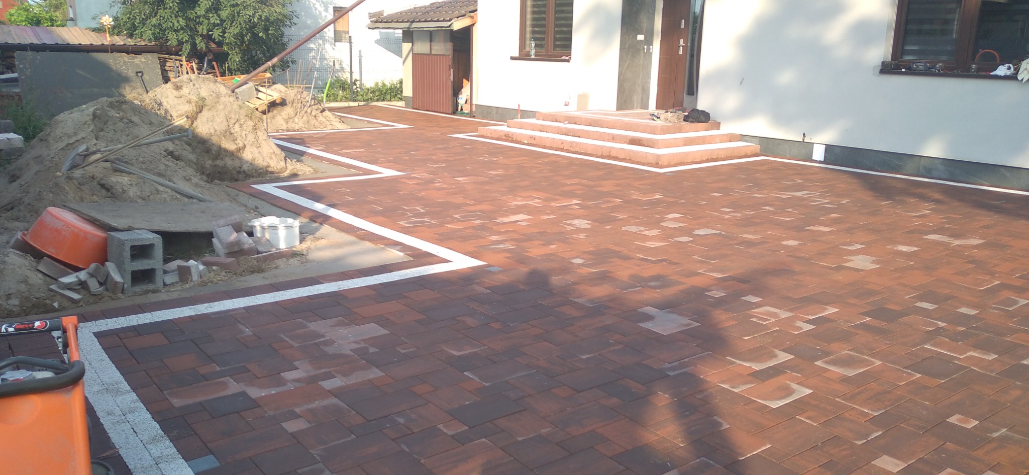 Ułożona kostka brukowa w odcieniach czerwieni z białymi obrzeżami prowadząca do schodów wejściowych domu, obok sterta piasku i narzędzia budowlane.