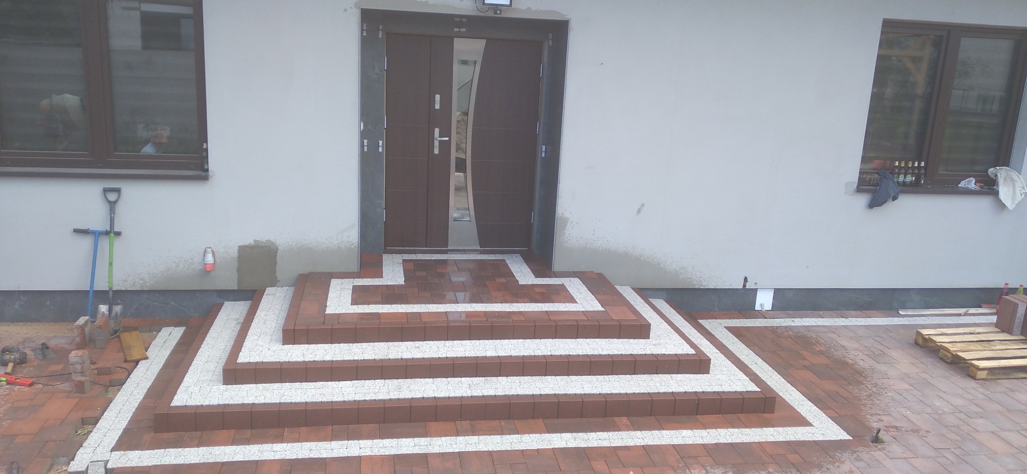 Schody wejściowe do domu wyłożone czerwoną kostką brukową z białymi akcentami, widoczne narzędzia brukarskie i paleta z kostką obok, okno z widocznymi butelkami w tle.