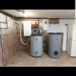 Dwa szare zbiorniki akumulacyjne ciepła marki Trinity, podłączone do białego pieca Hyundai z pomarańczowym naczyniem wzbiorczym, instalacja w kotłowni wyłożonej brązowymi płytkami.