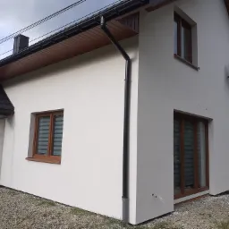 Świeżo otynkowana elewacja domu jednorodzinnego z widoczną czarną rynną, oknami z brązowymi ramami i anteną satelitarną, na tle zielonego krajobrazu.