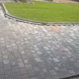 Ułożona szara kostka brukowa tworząca ścieżkę i patio w ogrodzie, z widocznym trawnikiem, obrzeżami i odpływem liniowym przy schodach.