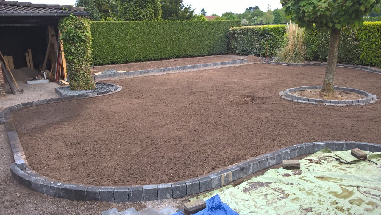 Przygotowany teren pod trawnik z obrzeżami z kostki granitowej, widoczny pień drzewa z obłożonymi kamieniami i zielony żywopłot w tle. Częściowo rozłożona plandeka ochronna.