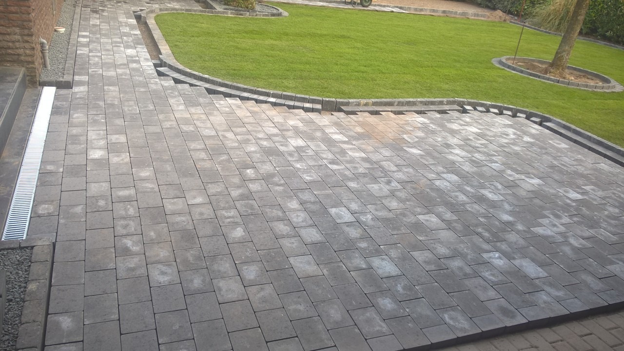 Ułożona szara kostka brukowa tworząca ścieżkę i patio w ogrodzie, z widocznym trawnikiem, obrzeżami i odpływem liniowym przy schodach.