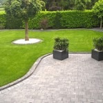 Uporządkowany ogród z równo przystrzyżonym trawnikiem, drzewkiem formowanym w kulę na trawniku otoczonym białym kamieniem, ścieżką z kostki brukowej z obrzeżem oraz dwoma donicami z krzewami. W tle...