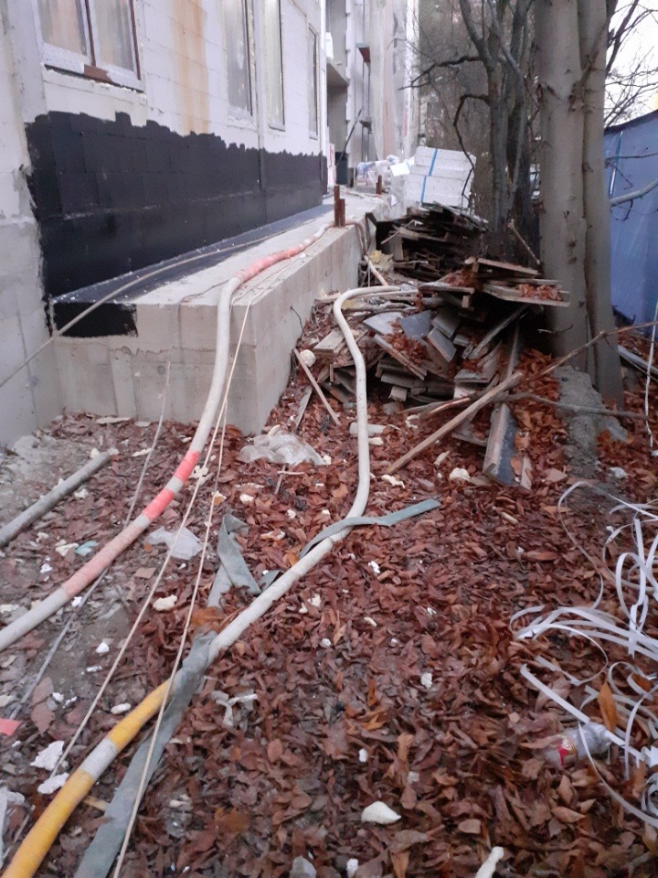 Stos płyt i odpadów drewnianych leżący na ziemi pokrytej liśćmi, obok fundamentów nowo budowanego budynku z widoczną izolacją przeciwwilgociową.