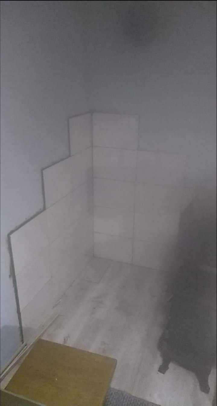 Fragment łazienki w trakcie układania glazury, widoczne częściowo ułożone białe płytki na ścianie, drewnopodobna podłoga i fragment taboretu w rogu.