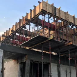 Wznoszenie dwukondygnacyjnego domu z betonu komórkowego z drewnianym szalunkiem stropu i widocznymi podporami stalowymi, w tle błękitne niebo z księżycem.