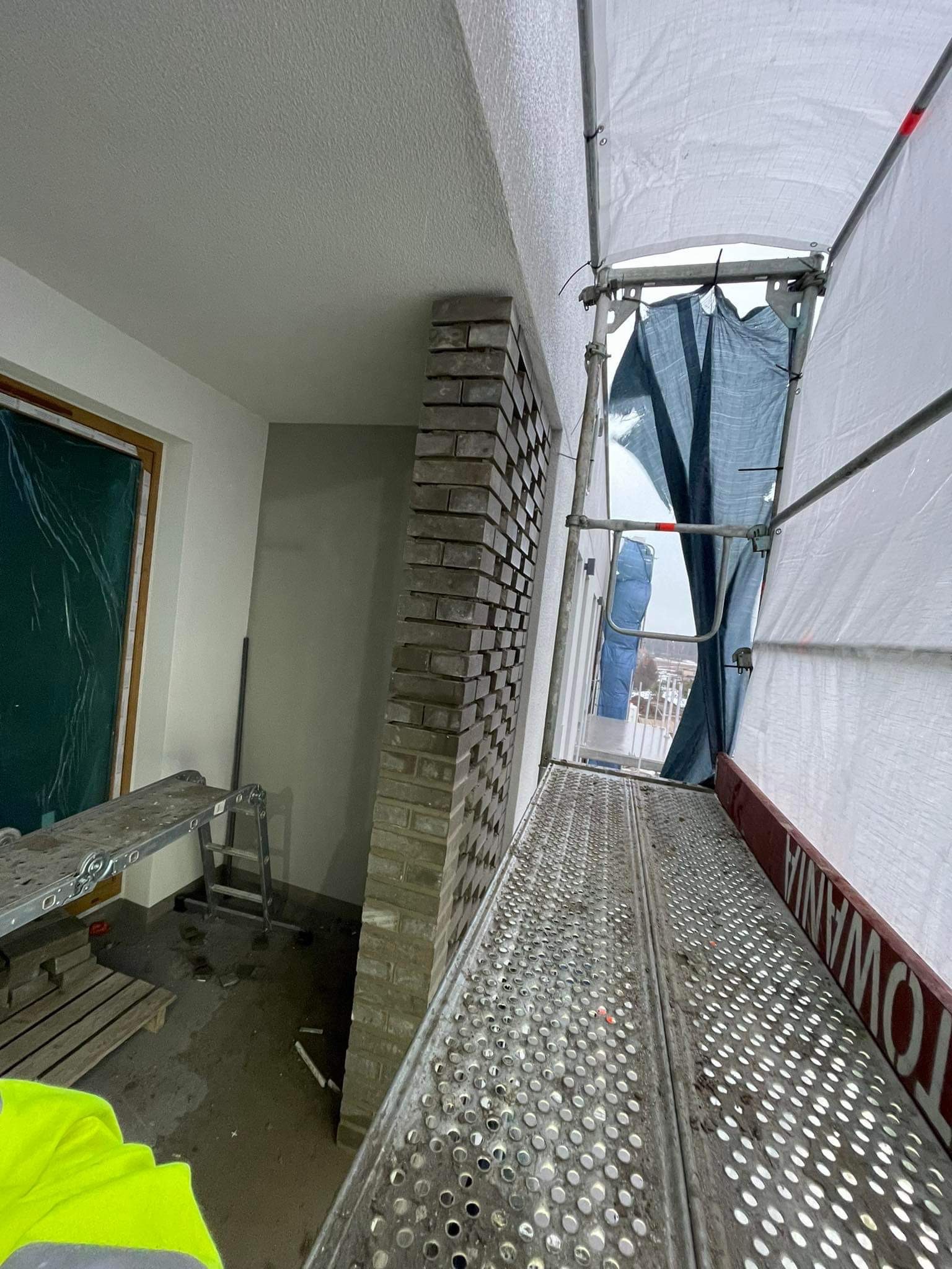Wewnętrzna ściana z cegły w trakcie murowania, widoczna ze rusztowania, okno zabezpieczone folią.