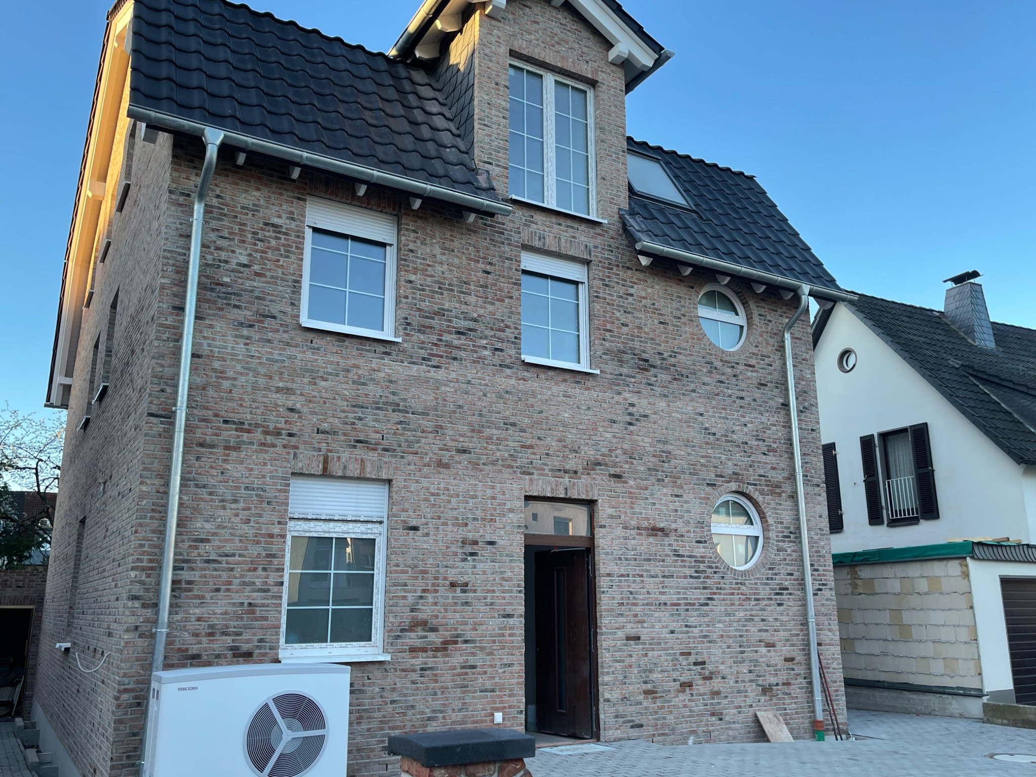 Nowoczesny, dwukondygnacyjny dom z cegły klinkierowej z czarną dachówką, okrągłymi oknami i pompą ciepła na zewnątrz.