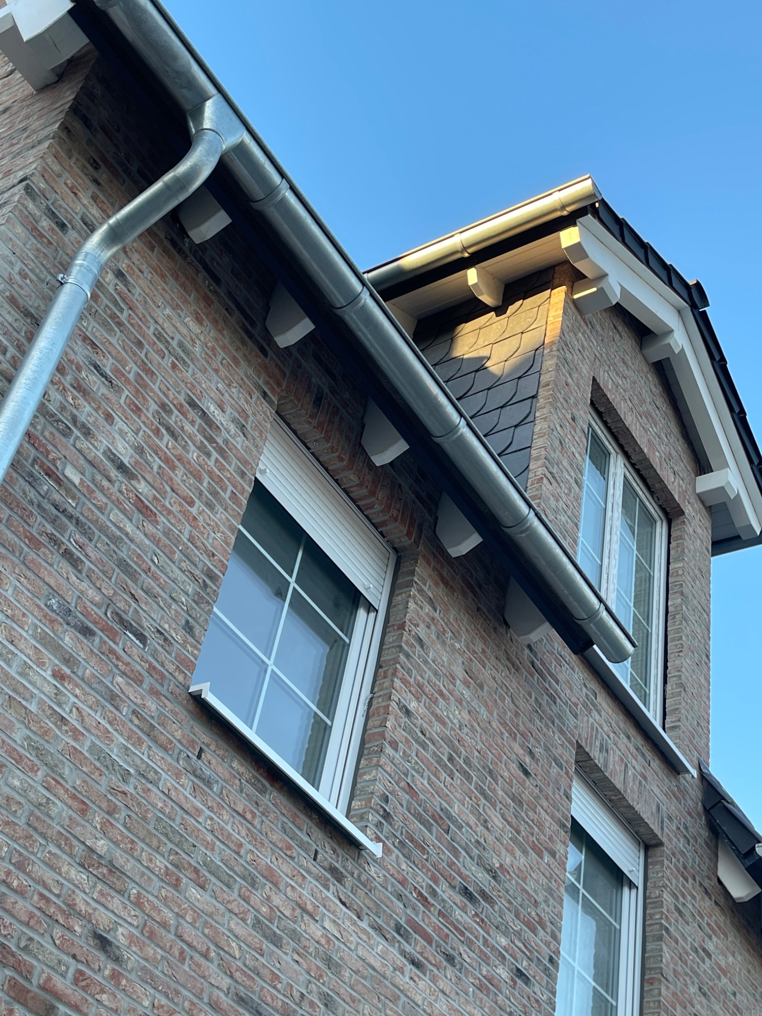 Fragment murowanego domu z cegły z widocznymi oknami z roletami zewnętrznymi, orynnowaniem i fragmentem dachu pokrytego dachówką w promieniach zachodzącego słońca.