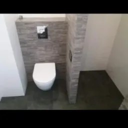 Nowoczesna toaleta podwieszana z szarą mozaiką kamienną na ścianie, ciemna podłoga, metalowy przycisk spłukujący.