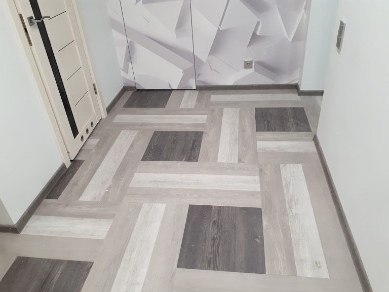 Wzór podłogi z paneli w odcieniach szarości i bieli ułożonych w kwadraty i prostokąty, widoczne drzwi z czarnymi wstawkami i ściana z geometrycznym wzorem w tle.