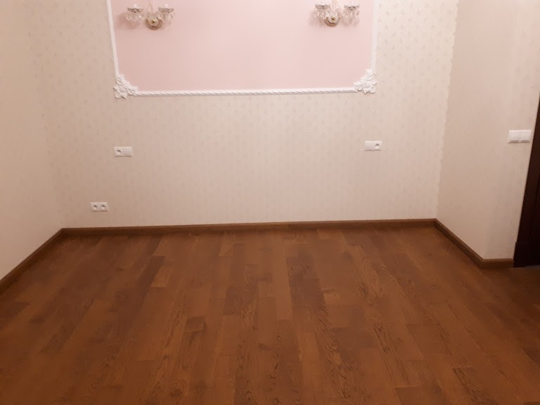 Wnętrze pokoju z nową, ciemnobrązową podłogą z desek, jasne ściany z dekoracyjnym panelem i dwoma kinkietami w górnej części ściany, widoczny fragment ciemnych drzwi po prawej stronie.