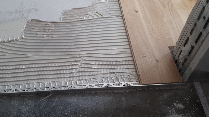 Układanie parkietu w Warszawie: widok z bliska na świeżo nałożony klej z charakterystycznymi rowkami, metalową listwę i fragment ułożonej deski podłogowej przy ścianie.