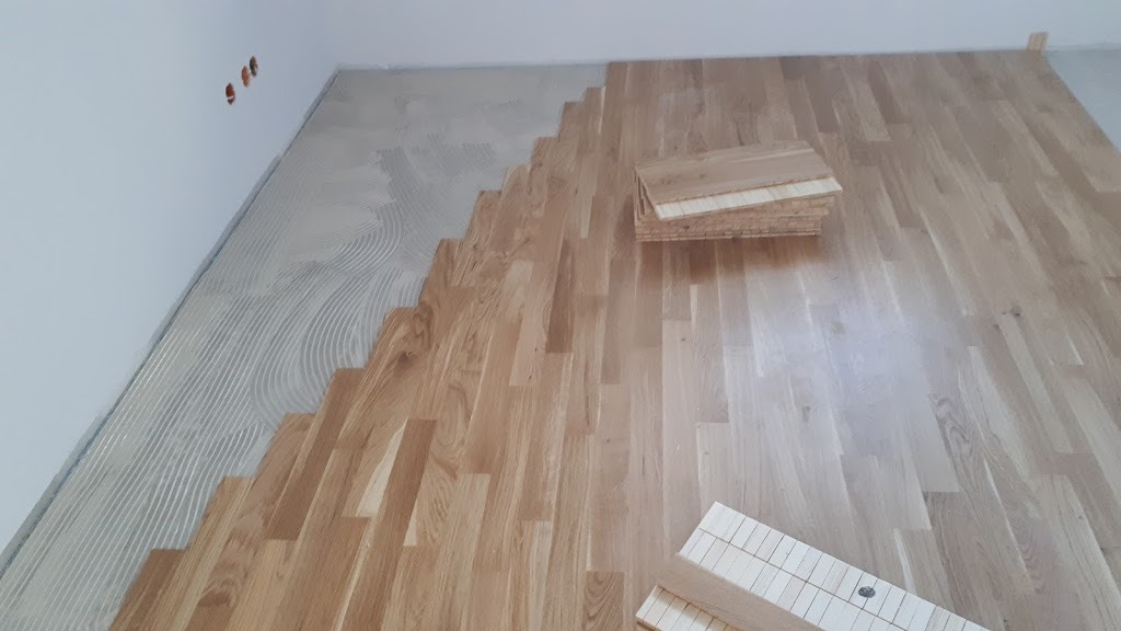 Fragment podłogi z jasnego drewna w trakcie układania parkietu; widoczny klej na podłożu i stos desek parkietowych.