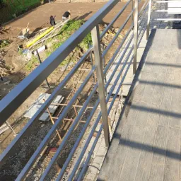 Szara, metalowa balustrada z poziomymi prętami widziana z góry, w tle prace ziemne i osoby przy nich pracujące.