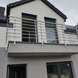 Nowoczesna balustrada balkonowa ze stali nierdzewnej, z poziomymi prętami, zamontowana na balkonie budynku w odcieniach szarości i bieli, z widocznymi oknami z roletami.