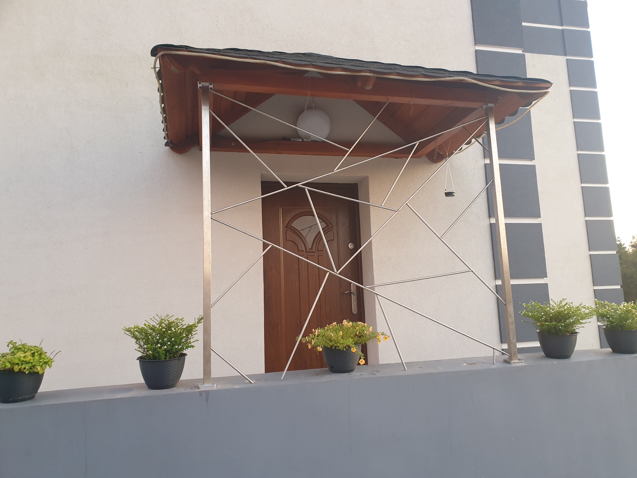 Nowoczesne zadaszenie nad drzwiami wejściowymi z geometryczną konstrukcją ze stali nierdzewnej, cztery donice z roślinami na podeście.