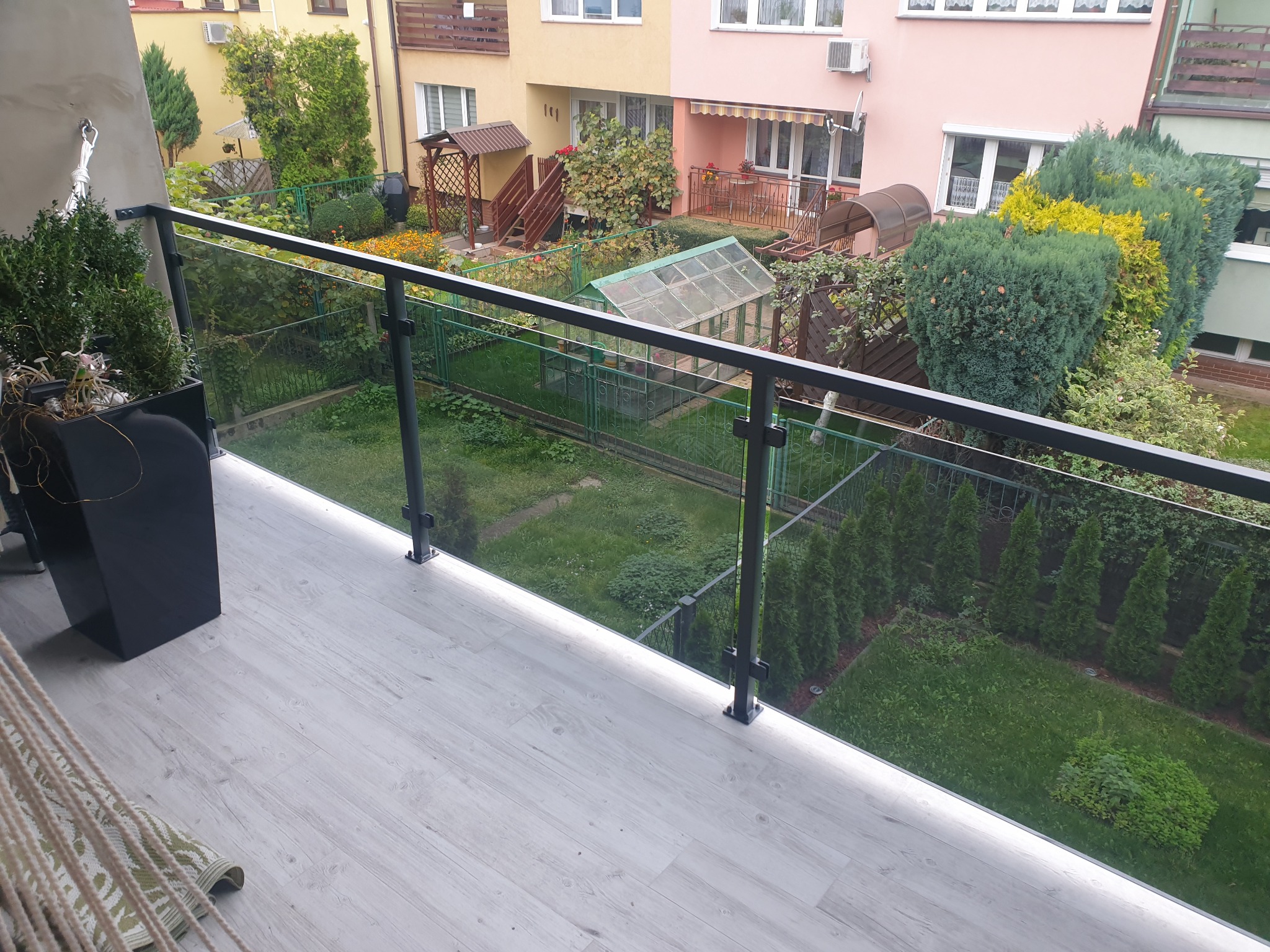 Balkon z balustradą szklaną z czarnymi elementami mocującymi, widok na zadbany ogród z zielonymi krzewami i budynkami w tle.