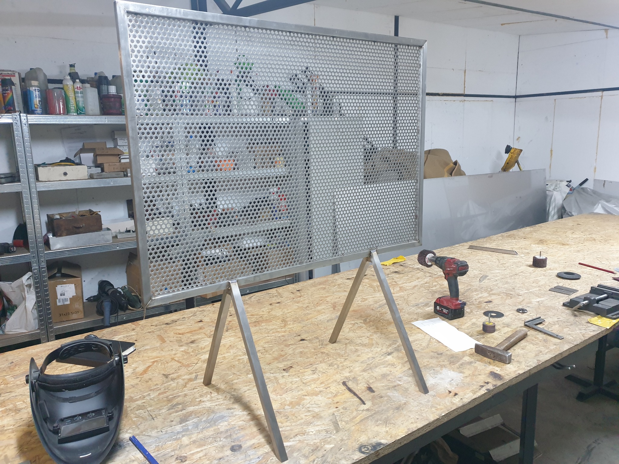 Metalowa perforowana płyta z ramą, ustawiona na metalowych podporach w warsztacie, obok leży maska spawalnicza i narzędzia.
