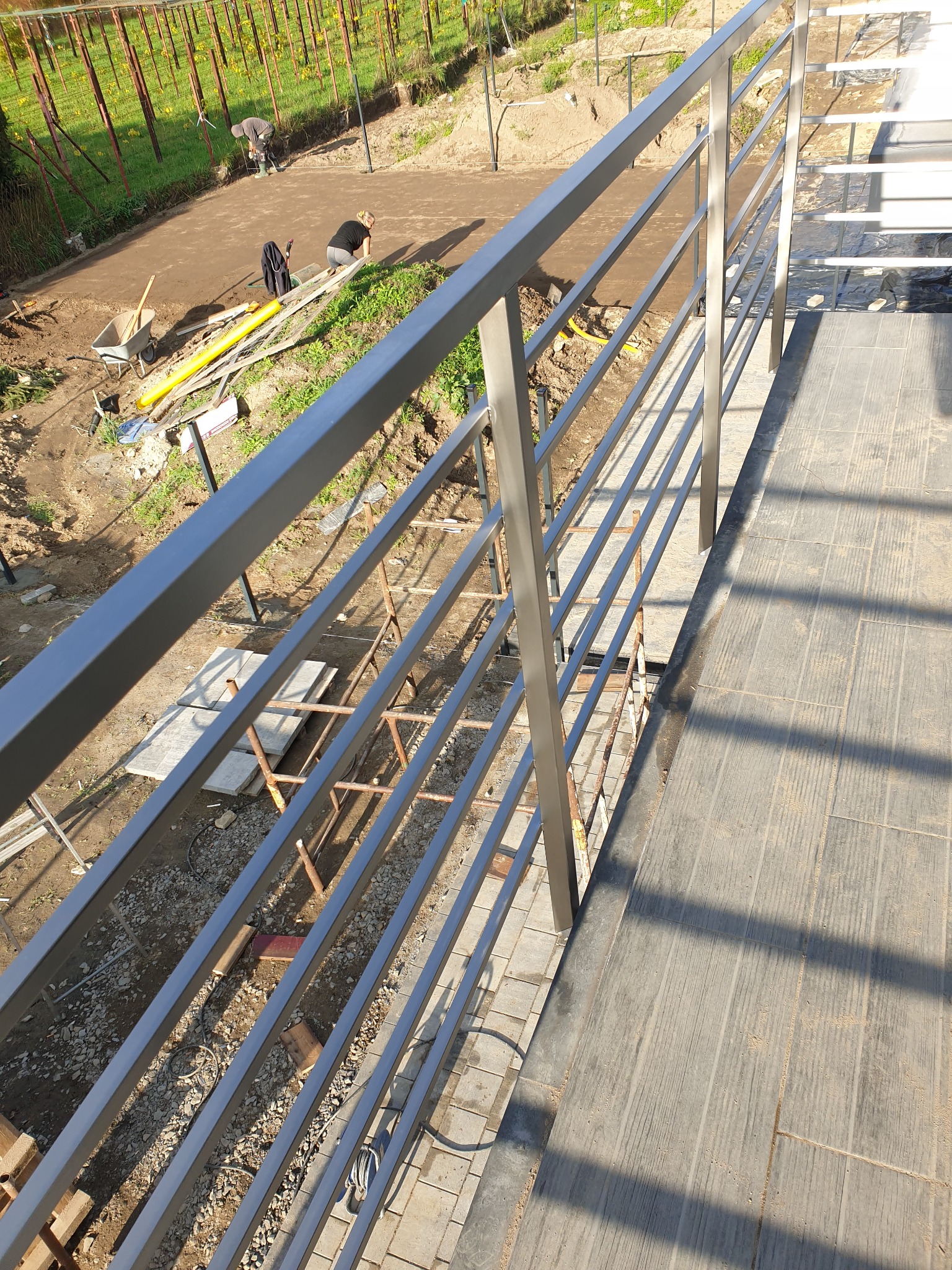 Szara, metalowa balustrada z poziomymi prętami widziana z góry, w tle prace ziemne i osoby przy nich pracujące.