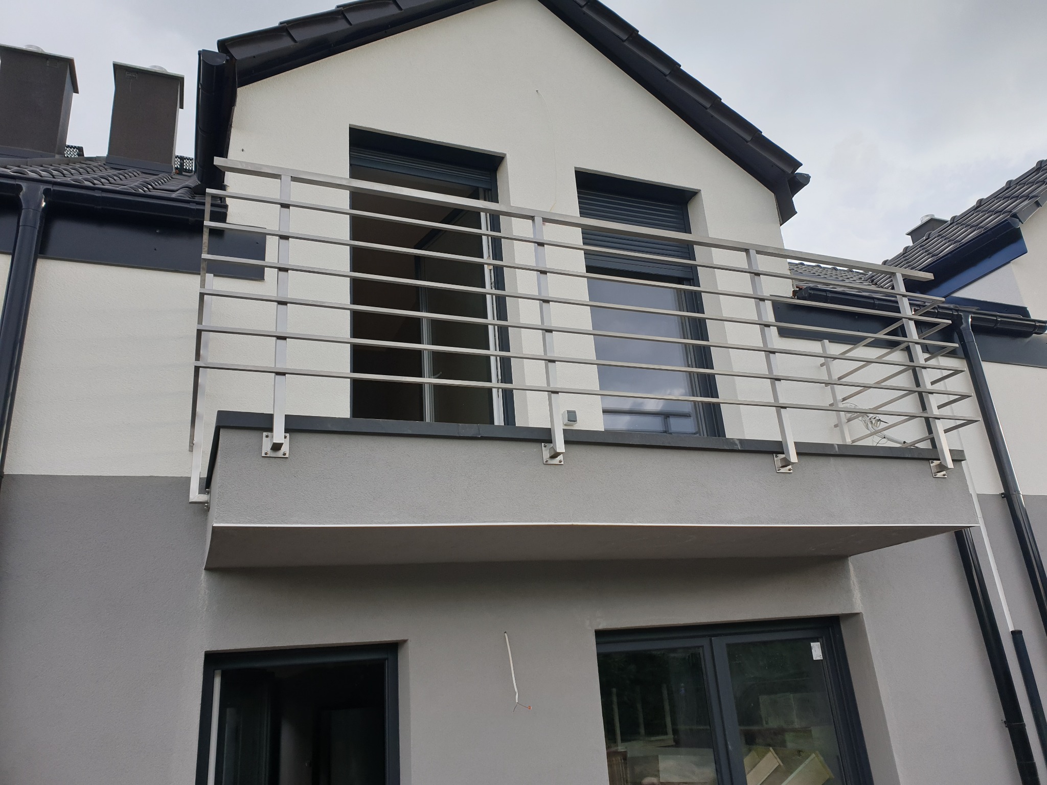 Nowoczesna balustrada balkonowa ze stali nierdzewnej, z poziomymi prętami, zamontowana na balkonie budynku w odcieniach szarości i bieli, z widocznymi oknami z roletami.