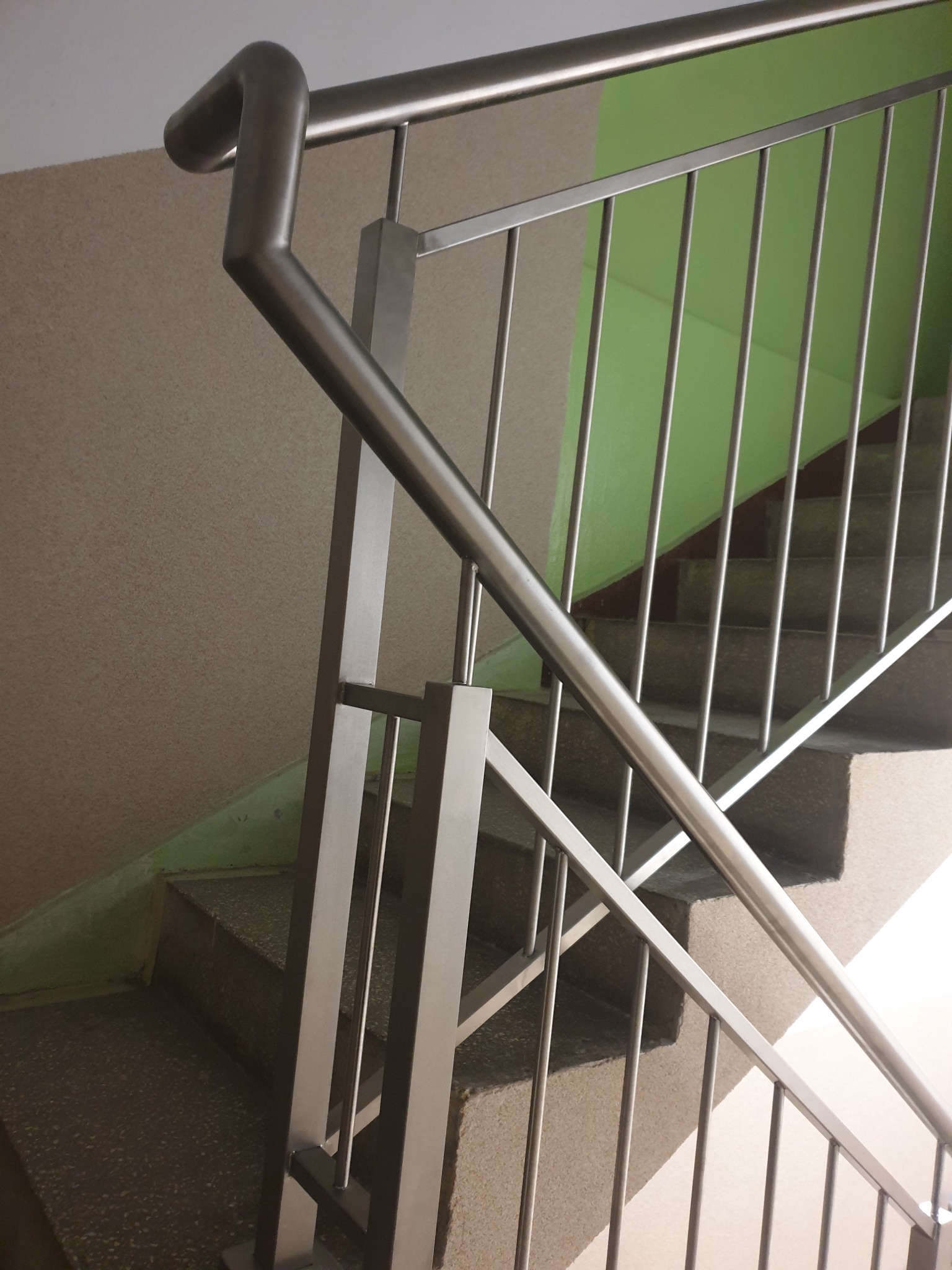 Wewnętrzna klatka schodowa z metalową balustradą z pionowymi prętami i zaokrąglonym poręczą, widok z dołu na schody z zieloną ścianą w tle.