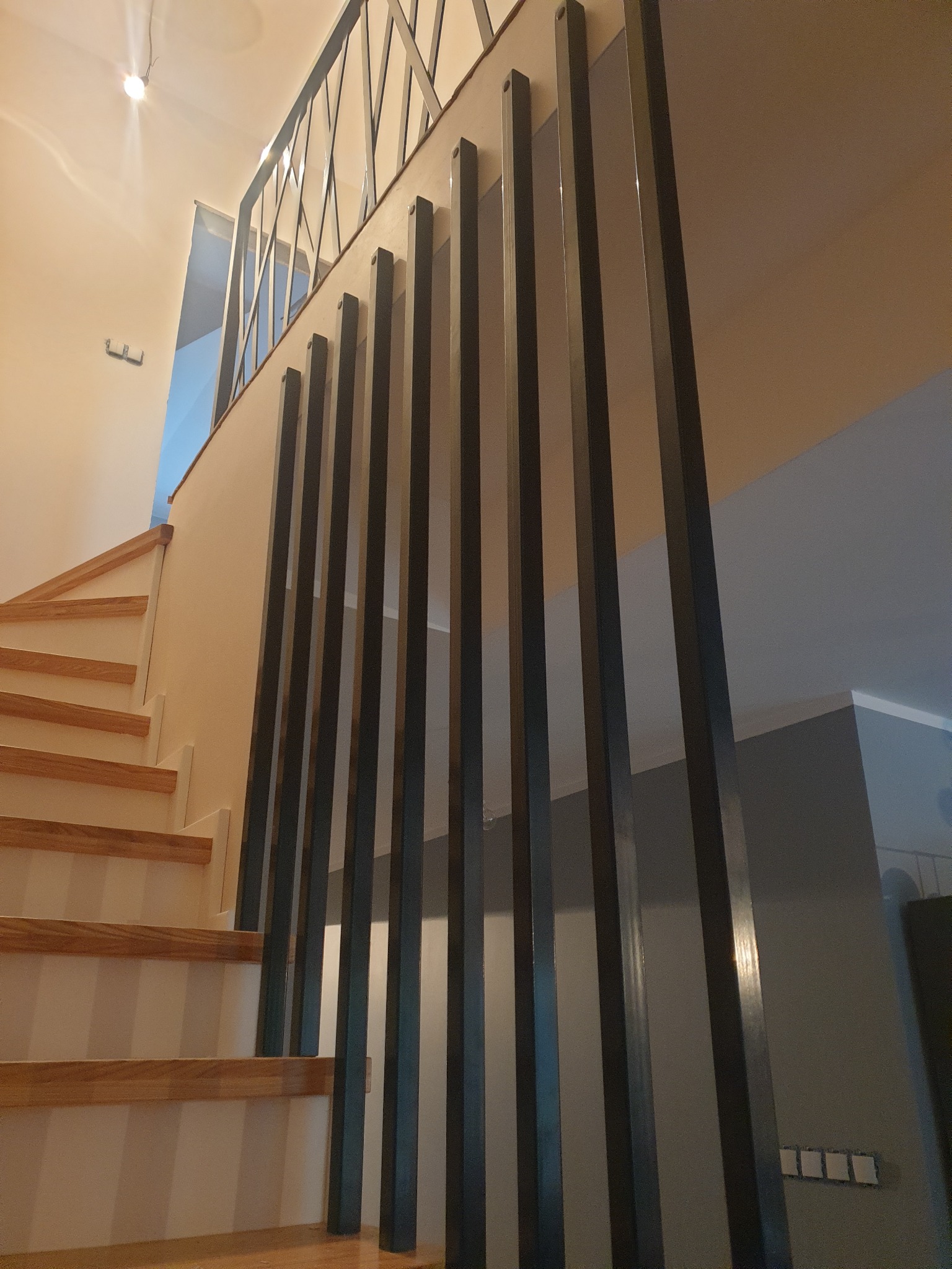 Nowoczesne schody z drewnianymi stopniami i balustradą z pionowych, ciemnych, metalowych prętów, widziane z dołu.