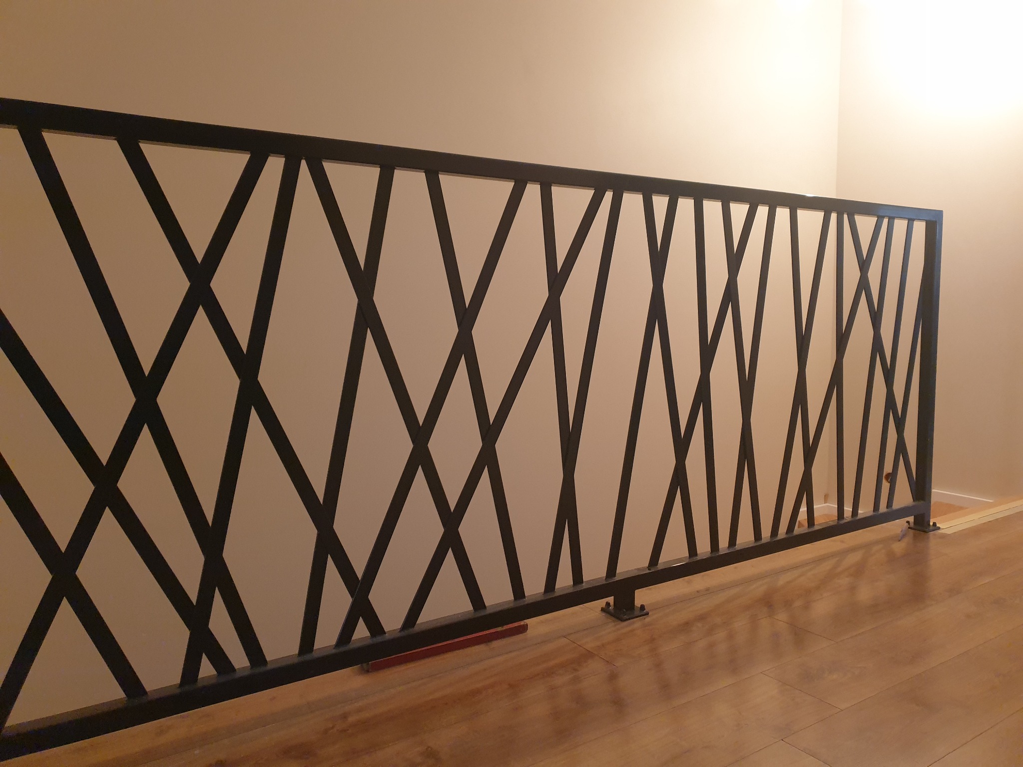 Czarna, metalowa barierka z geometrycznym wzorem krzyżujących się ukośnych prętów, zamontowana na drewnianej podłodze przy jasnej ścianie.