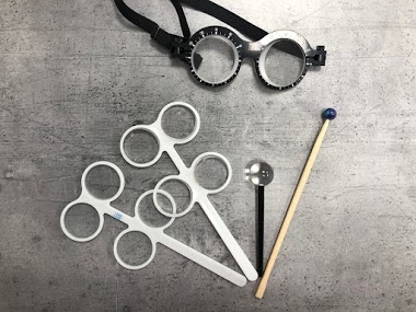 Zestaw narzędzi okulistycznych: okulary próbne z regulowanym rozstawem, trzy oprawki do soczewek próbnych, pręt z niebieską kulką i przyrząd z soczewką na szarym tle.