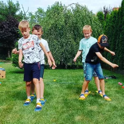 Czterech chłopców na trawniku bawi się w grę zespołową na drewnianych nartach, z zielonymi krzewami w tle.