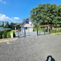 Nowoczesna posesja z szarym ogrodzeniem panelowym, bramą wjazdową i furtką, widok na domek letniskowy z szarą elewacją i drewnianą altaną w tle.