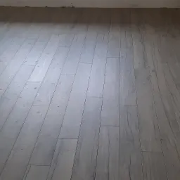 Ułożone, szare panele podłogowe imitujące drewno w trakcie prac wykończeniowych, widoczne wystające kable elektryczne ze ściany.