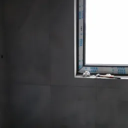 Szare płytki wielkoformatowe na ścianach i podłodze w surowym stanie, okno PCV z widocznym logo VEKA, młotek i szmatki na parapecie.