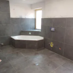 Niedokończona łazienka z szarymi płytkami, narożna wanna z obudową z płytek, widoczne wystające kable i instalacje hydrauliczne, okno w narożniku pomieszczenia z zabezpieczeniem folią.