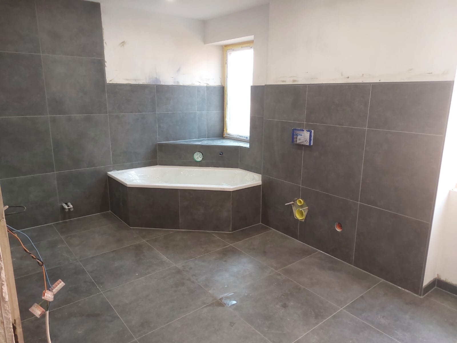 Niedokończona łazienka z szarymi płytkami, narożna wanna z obudową z płytek, widoczne wystające kable i instalacje hydrauliczne, okno w narożniku pomieszczenia z zabezpieczeniem folią.
