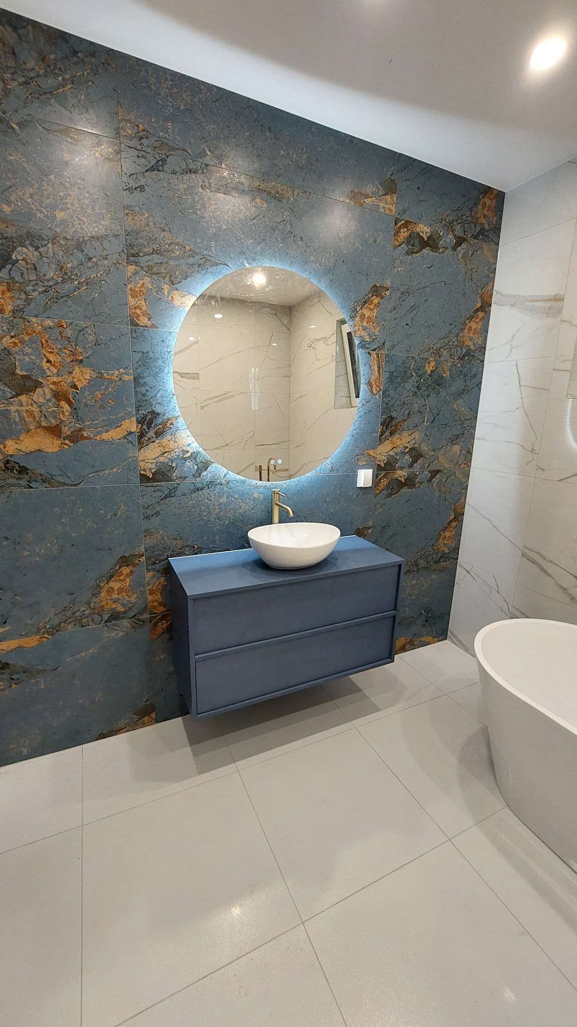 Niebieska szafka z umywalką i okrągłym lustrem z podświetleniem LED na tle ściany z płytek imitujących marmur w wykończonej łazience.