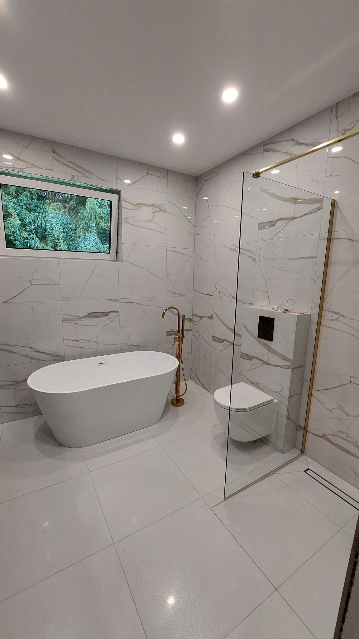 Elegancka łazienka z wolnostojącą wanną, złotą armaturą i kabiną prysznicową ze złotymi detalami. Marmurowe płytki na ścianach i podłodze. Okno z widokiem na zieleń.