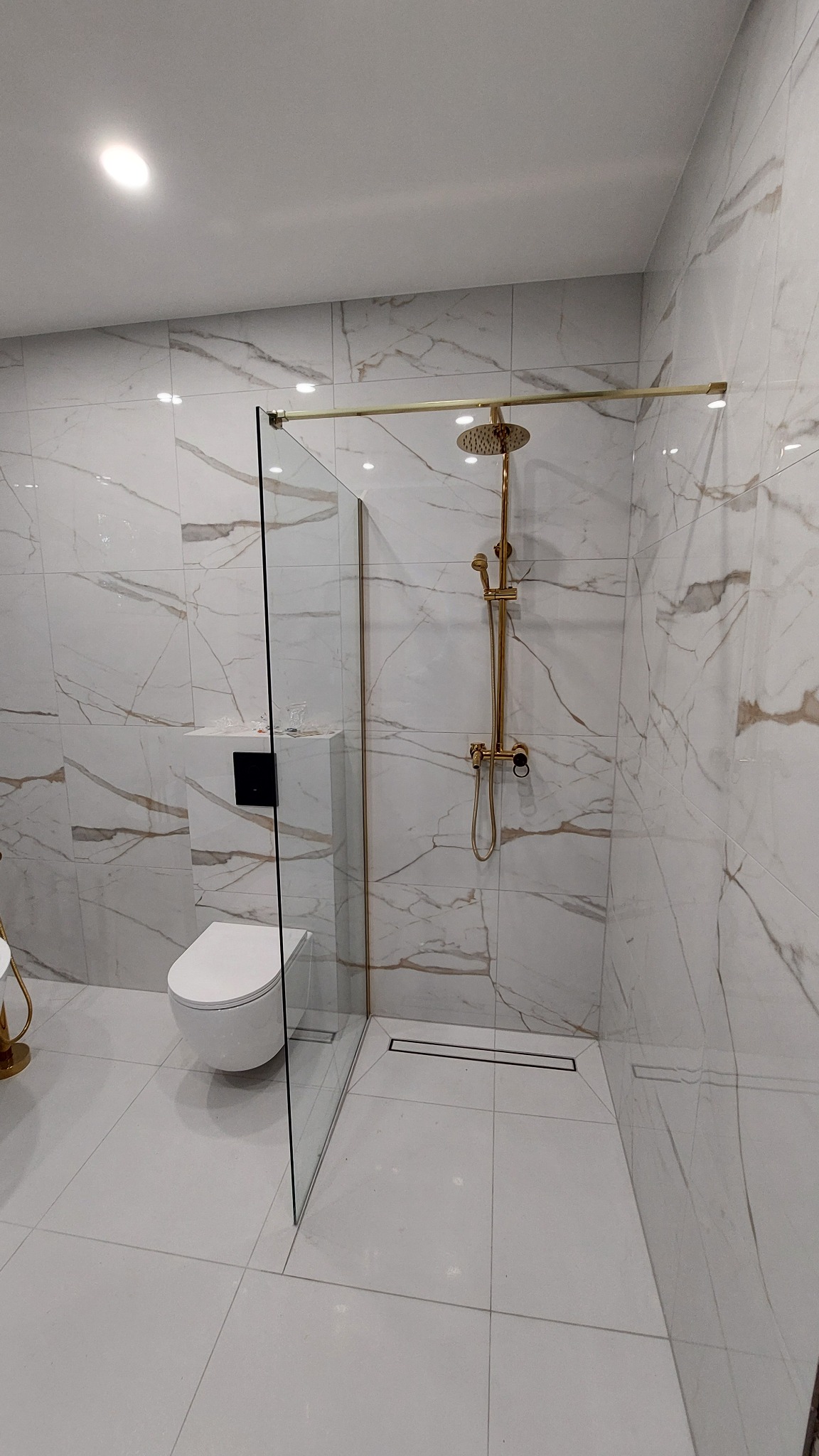 Nowoczesna łazienka z kabiną typu walk-in, wykończona białymi płytkami imitującymi marmur z złotymi akcentami armatury i profili. Minimalistyczny design.