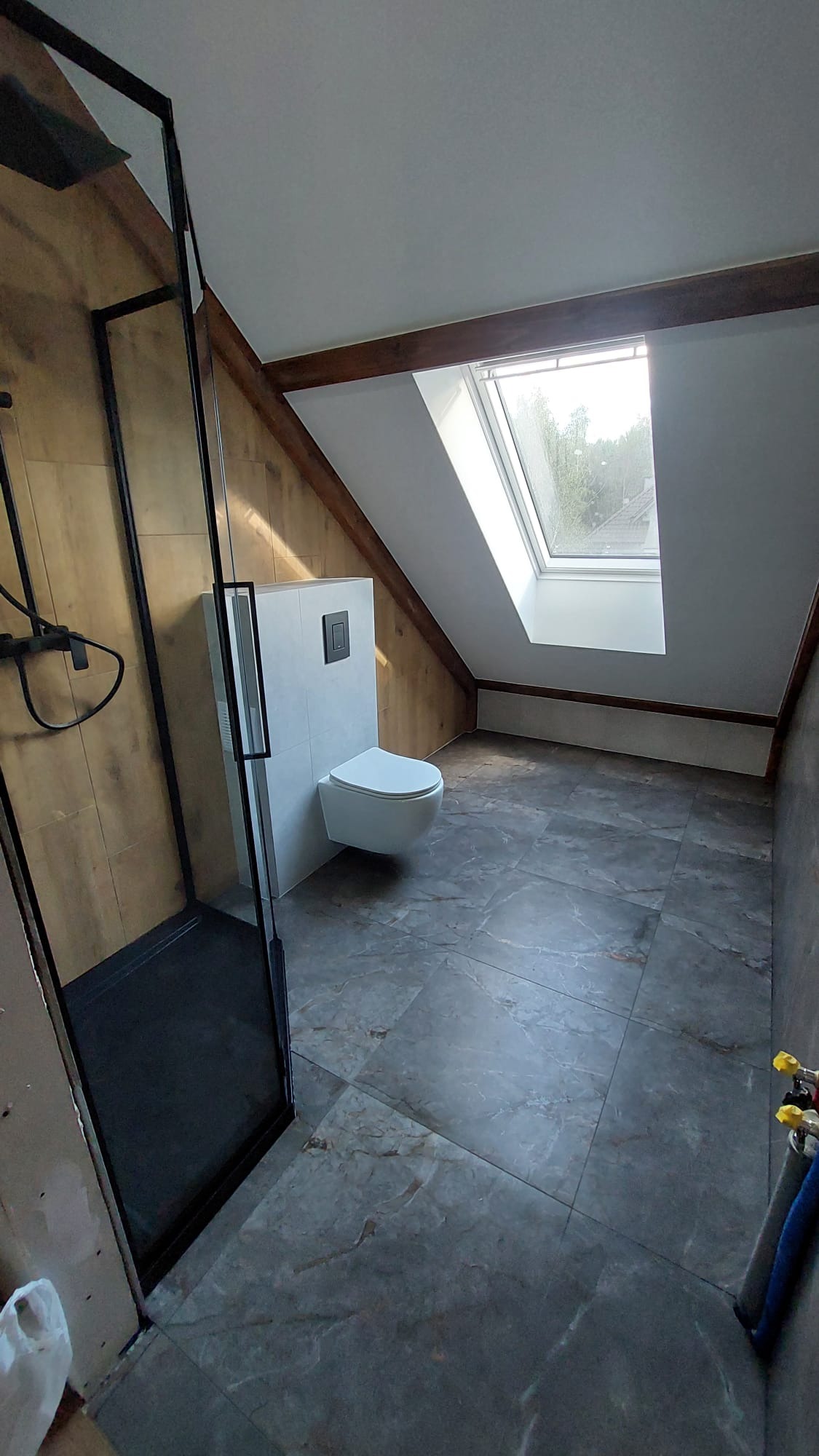 Wykończona łazienka na poddaszu ze skośnym sufitem i oknem dachowym. Kabina prysznicowa z czarną ramą, podwieszana toaleta i szare płytki podłogowe.