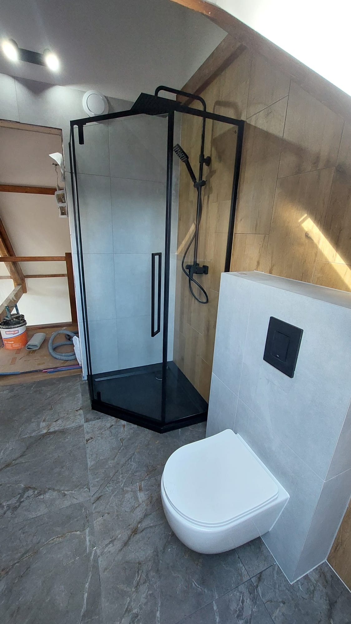 Nowoczesna kabina prysznicowa z czarnymi profilami i deszczownicą, obok podwieszana toaleta. Ukończona aranżacja łazienki na poddaszu z szarą marmurową podłogą.