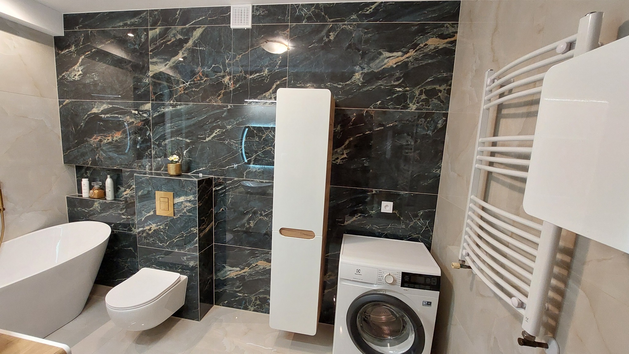 Elegancka łazienka z wanną wolnostojącą, toaletą podwieszaną, szafką i pralką. Ściany wyłożone płytkami imitującymi marmur. Grzejnik drabinkowy.