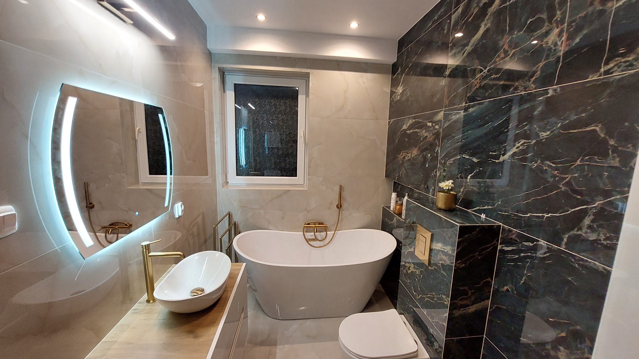 Elegancka łazienka z wolnostojącą wanną, umywalką nablatową i podświetlanym lustrem. Marmurowe płytki w odcieniach beżu i czerni dodają luksusowego charakteru.