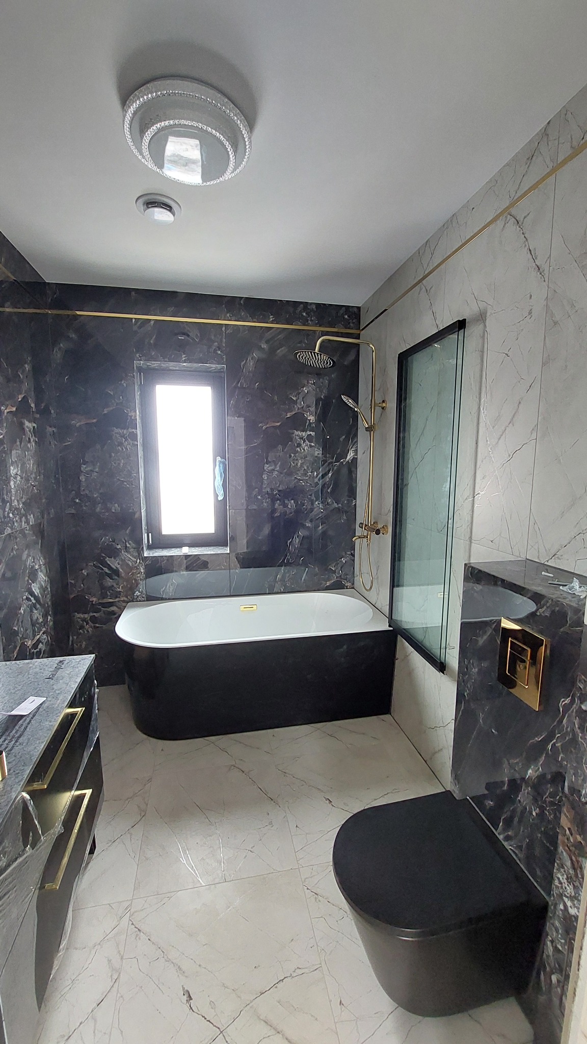 Elegancka łazienka w czarno-białej kolorystyce z marmurowymi płytkami, złotymi akcentami i nowoczesną armaturą. Widoczna wanna, toaleta i umywalka.