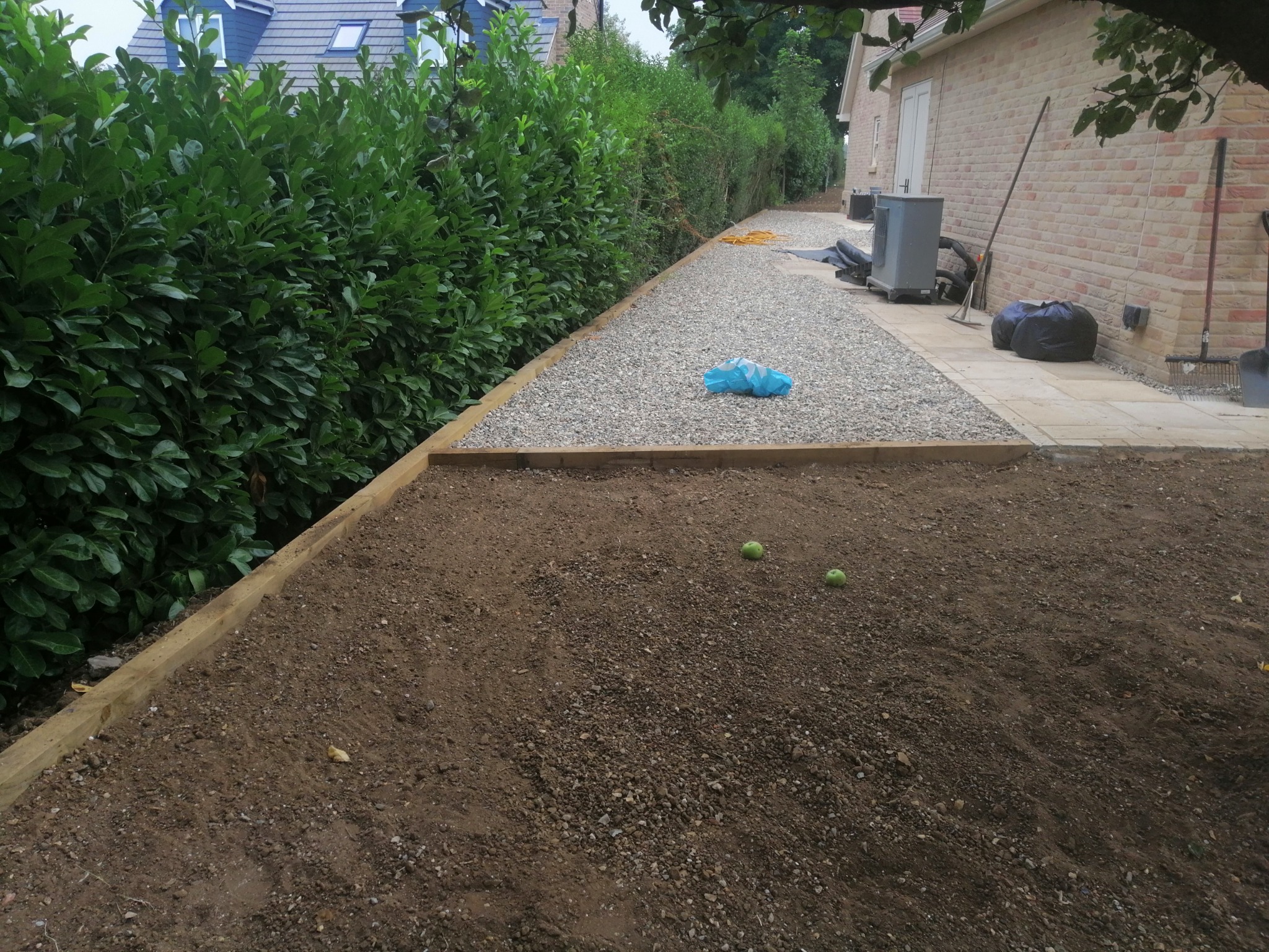 Przygotowanie terenu pod ogród: wyrównana ziemia z drewnianym obramowaniem, obok ścieżka wysypana żwirem, w tle żywopłot i budynek.