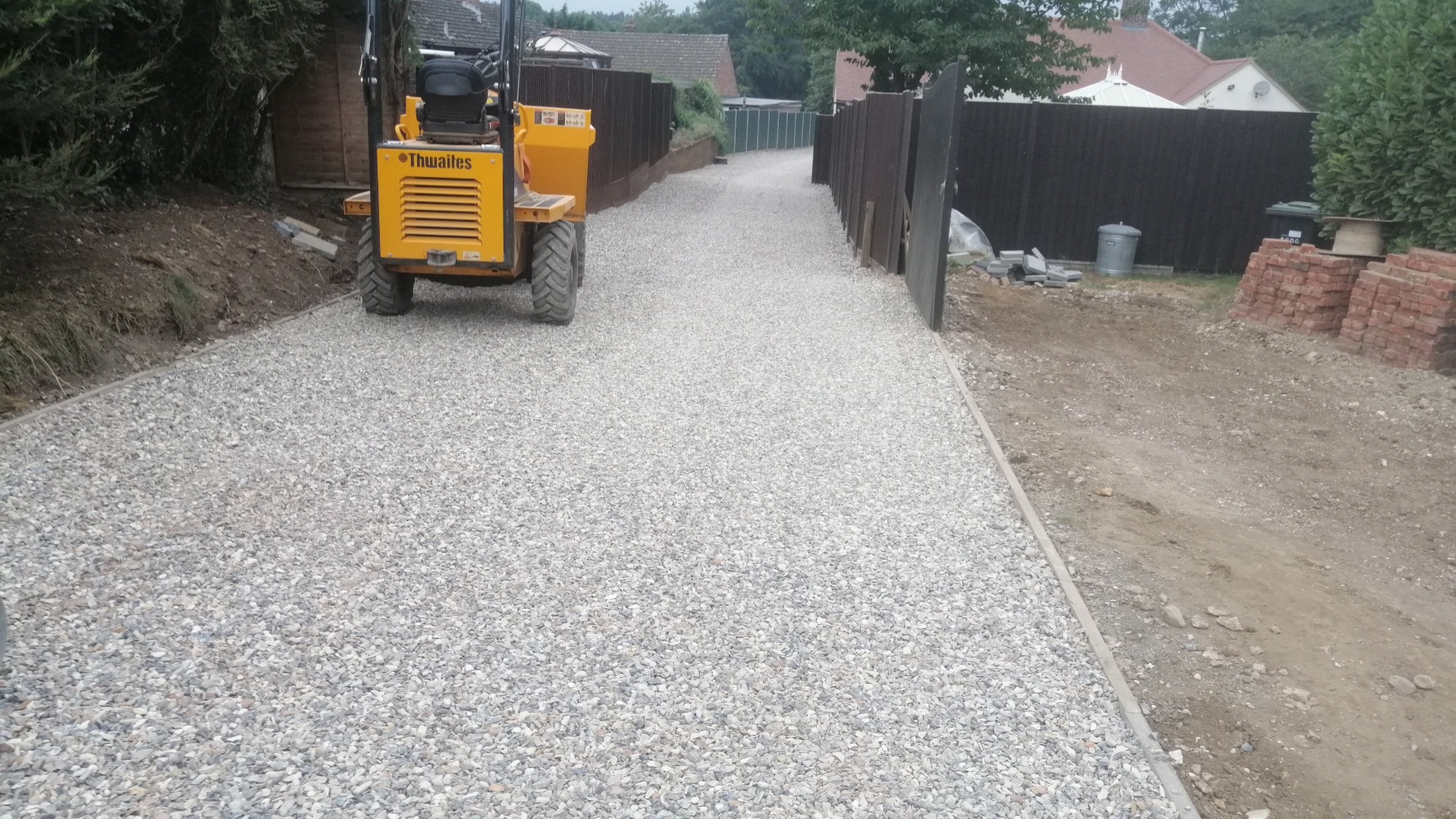Żwirowy podjazd z obrzeżami, widoczny mały wywrotka Thwaites i częściowo zamontowane ogrodzenie z ciemnego drewna.