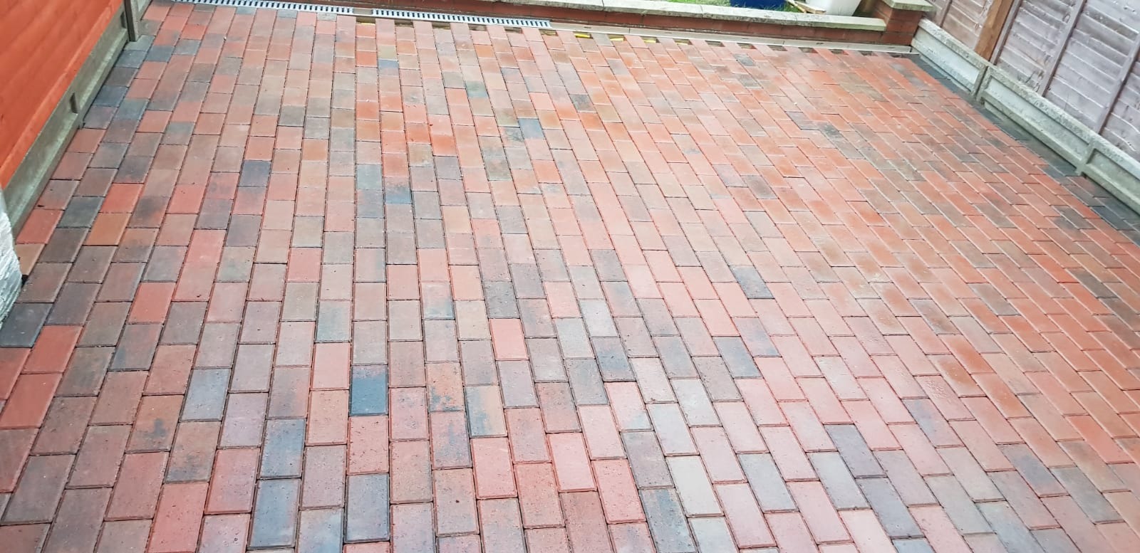 Ułożona, czerwono-szara kostka brukowa na podjeździe z widocznym odpływem liniowym i fragmentami ogrodzenia oraz budynku.