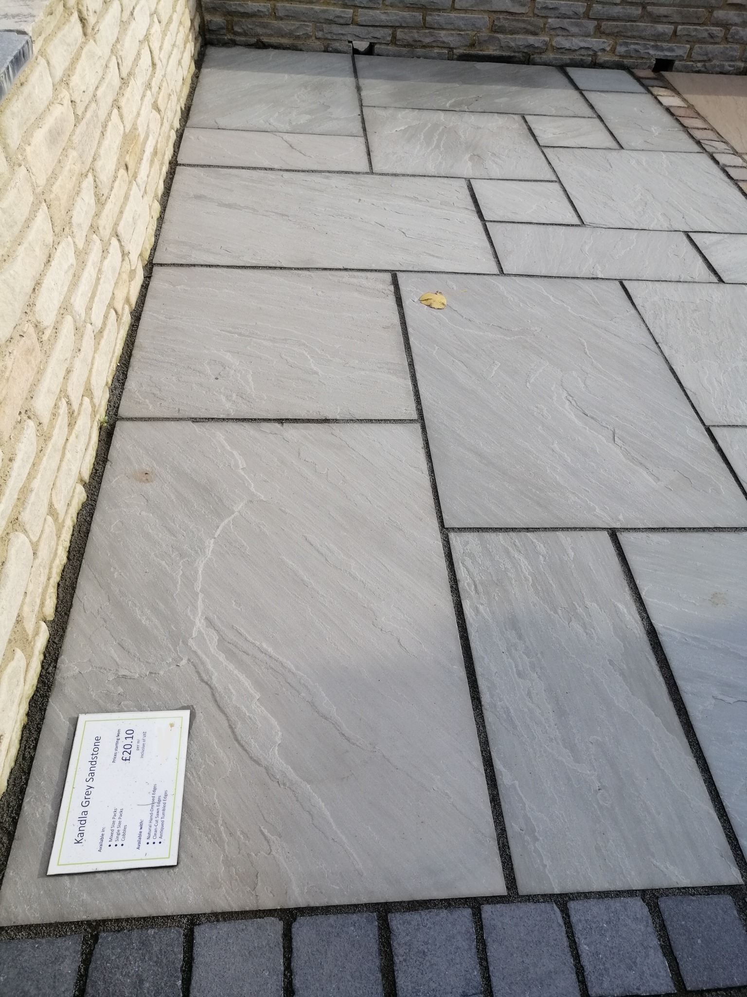 Świeżo ułożona nawierzchnia z szarych płyt piaskowca 'Kandla Grey Sandstone' z widoczną metką informacyjną, ułożona w nieregularny wzór, obramowana ciemnoszarą kostką i jasną ścianą z kamienia.