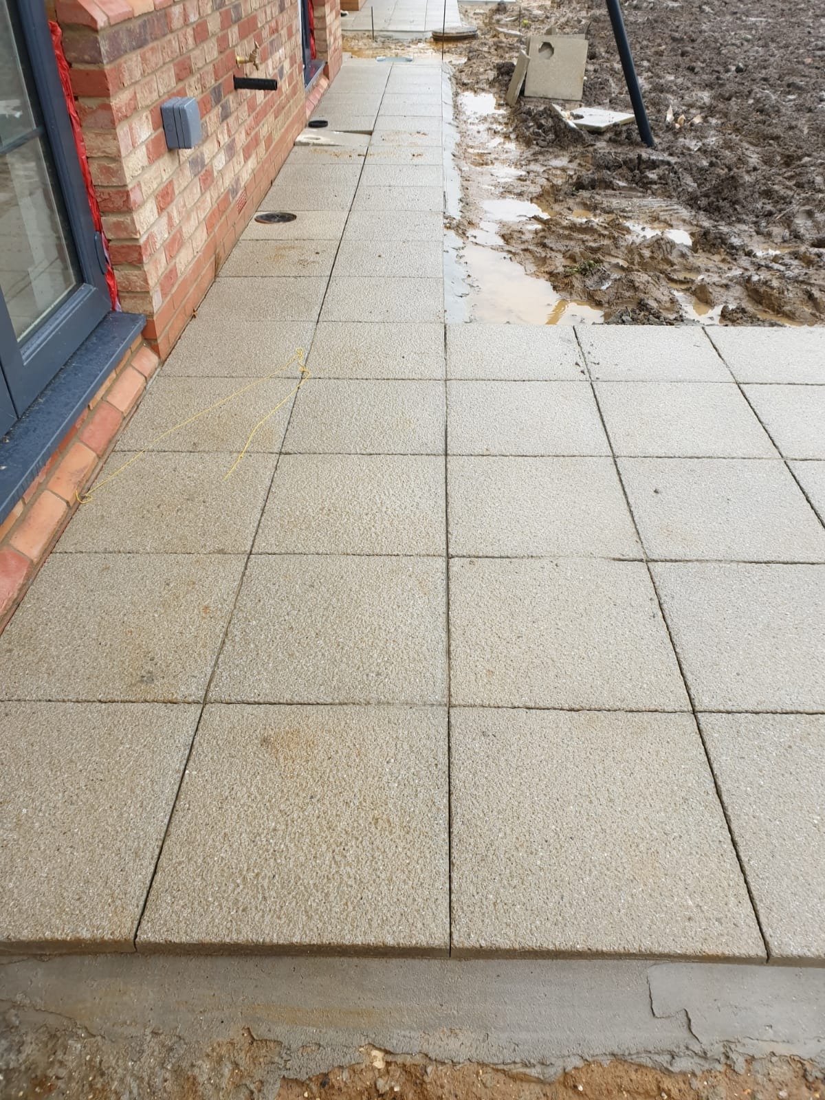 Świeżo ułożony chodnik z jasnoszarej kostki brukowej kwadratowej, przylegający do ściany budynku z cegły, z widocznymi żółtymi sznurkami wyznaczającymi linię, oraz błotnistym terenem w tle.
